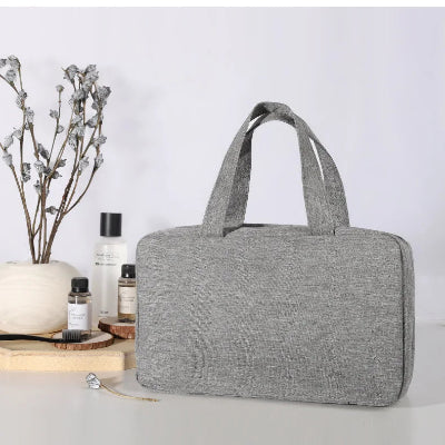 Trousse de toilette suspendue  Voyage Pratique gris fermée posée sur table blanche côté fleurs flacons parfums